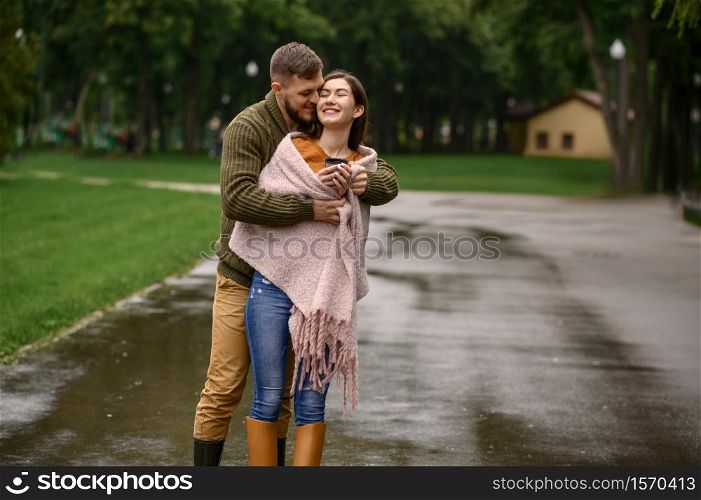 Love couple bask against each other in park, summer rainy day. Man and woman stand under umbrella in rain, romantic date on walking path, wet weather in alley. Love couple bask against each other in rainy day