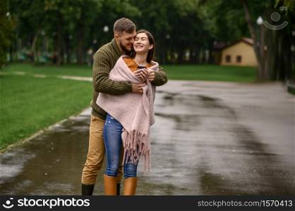 Love couple bask against each other in park, summer rainy day. Man and woman stand under umbrella in rain, romantic date on walking path, wet weather in alley. Love couple bask against each other in rainy day