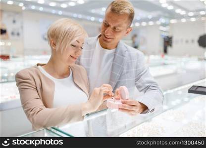 Love couple buying gold decoration. Male and female consumers looking on jewels in jewelry store. Man and woman choosing wedding rings. Future bride and groom in jewellery shop