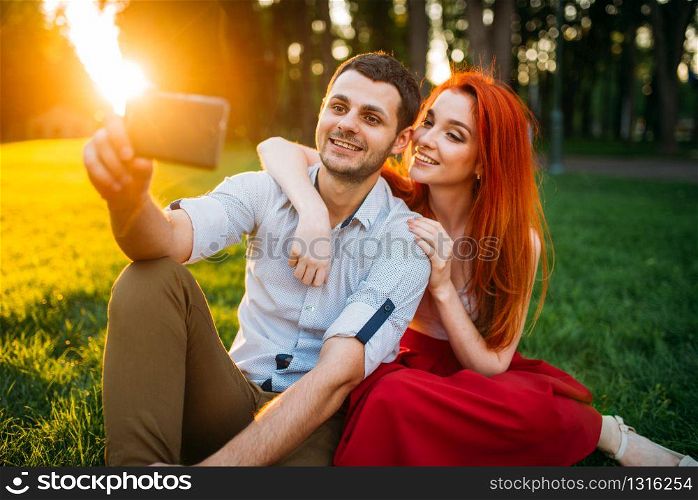 Love couple makes selfie on phone camera in summer park on sunset. Romantic date of attractive woman and young man. Love couple makes selfie in summer park on sunset