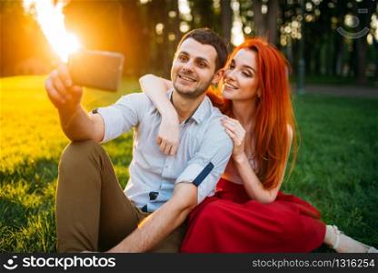Love couple makes selfie on phone camera in summer park on sunset. Romantic date of attractive woman and young man. Love couple makes selfie in summer park on sunset