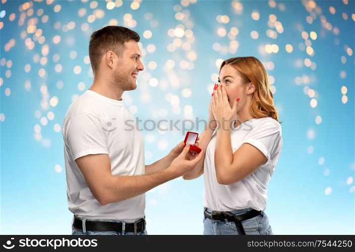 love, couple, proposal and holiday concept - man giving diamond engagement ring in little red box to happy woman over holidays lights on blue background. man giving woman engagement ring on valentines day