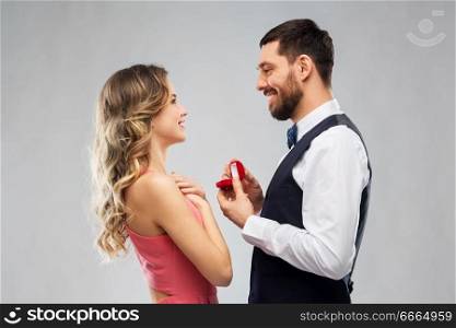 love, couple, proposal and people concept - happy man giving diamond engagement ring in little red box to woman over grey background. man giving woman engagement ring on valentines day