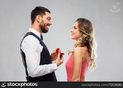 love, couple, proposal and people concept - happy man giving diamond engagement ring in little red box to woman over grey background. man giving woman engagement ring on valentines day