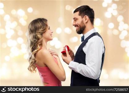 love, couple, proposal and people concept - happy man giving diamond engagement ring in little red box to woman over beige background with festive lights. man giving woman engagement ring on valentines day