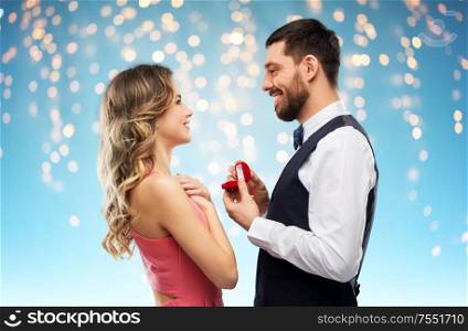 love, couple, proposal and people concept - happy man giving diamond engagement ring in little red box to woman over holiday lights on blue background. man giving woman engagement ring on valentines day