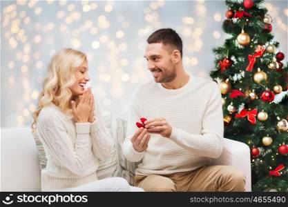 love, couple, proposal and people concept - happy man giving engagement ring in little red box to woman over christmas tree and lights background