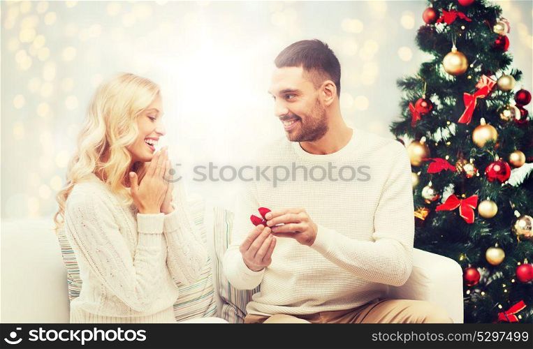 love, couple, proposal and people concept - happy man giving engagement ring in little red box to woman over christmas tree and lights background. man giving woman engagement ring for christmas