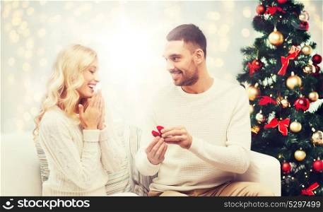 love, couple, proposal and people concept - happy man giving engagement ring in little red box to woman over christmas tree and lights background. man giving woman engagement ring for christmas
