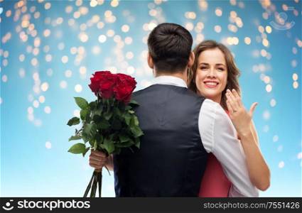 love, couple, proposal and people concept - happy woman with engagement ring and bunch of roses hugging man over holiday lights on blue background. woman with engagement ring and roses hugging man