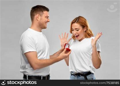 love, couple, proposal and people concept - man giving diamond engagement ring in little red box to happy woman over grey background. man giving woman engagement ring on valentines day