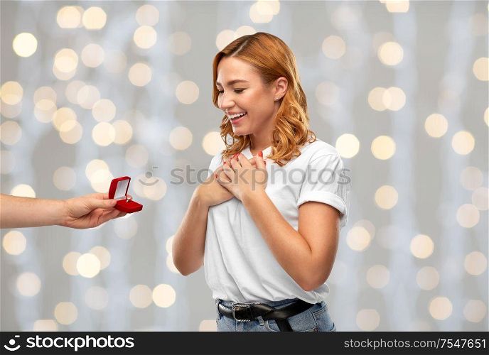 love, couple, proposal and people concept - man giving diamond engagement ring in little red box to happy woman over festive lights background. man giving woman engagement ring over lights