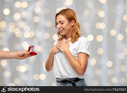 love, couple, proposal and people concept - man giving diamond engagement ring in little red box to happy woman over festive lights background. man giving woman engagement ring over lights