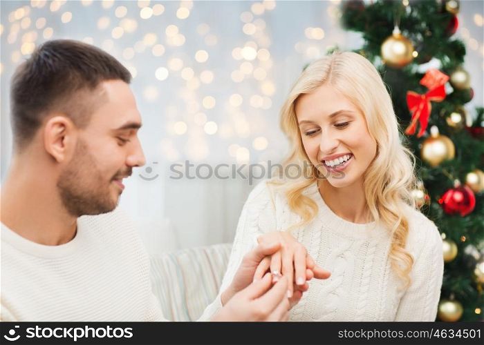 love, couple, proposal, holidays and people concept - happy man giving diamond engagement ring to woman over christmas tree and lights background