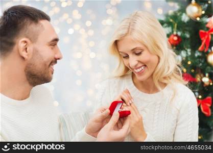 love, couple, proposal, holidays and people concept - happy man giving diamond engagement ring in little red box to woman over christmas tree and lights background