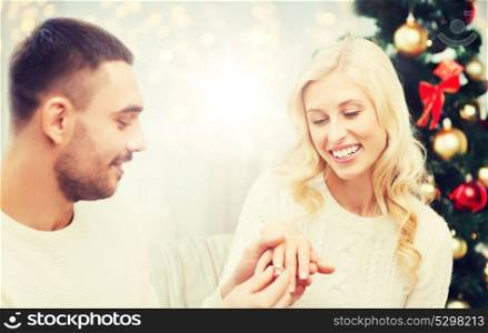 love, couple, proposal, holidays and people concept - happy man giving diamond engagement ring to woman over christmas tree and lights background. man giving woman engagement ring for christmas
