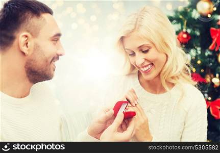 love, couple, proposal, holidays and people concept - happy man giving diamond engagement ring in little red box to woman over christmas tree and lights background. man giving woman engagement ring for christmas