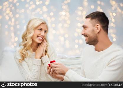 love, couple, relationship, proposal and holidays concept - happy man giving engagement ring in little red gift box to woman over christmas tree and lights background