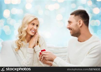 love, couple, relationship, proposal and holidays concept - happy man giving engagement ring in little red gift box to woman over blue holidays lights background