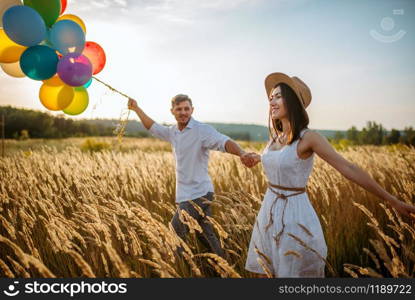 Love couple with balloons, leisure in a rye field on sunset. Pretty wife and husband walking on summer meadow. Love couple with balloons, leisure in a rye field