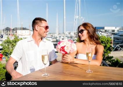 love, dating, happiness and people concept - smiling couple wearing sunglasses with bunch of flowers and champagne glasses looking to each other at cafe