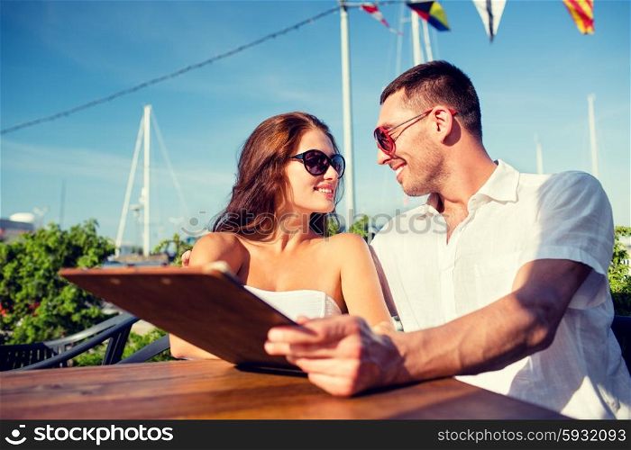 love, dating, people and food concept - smiling couple wearing sunglasses holding menu and looking to each other at cafe