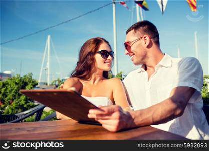 love, dating, people and food concept - smiling couple wearing sunglasses holding menu and looking to each other at cafe