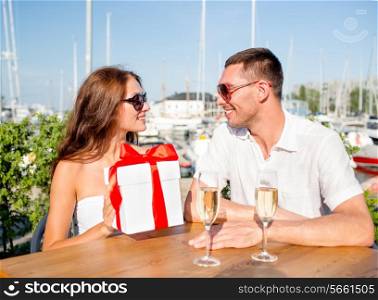 love, dating, people and holidays concept - smiling couple wearing sunglasses sitting with gift box, flowers and champagne at cafe