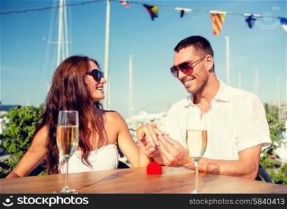 love, dating, people and holidays concept - smiling couple wearing sunglasses with champagne and small red gift box putting wedding ring on finger at cafe