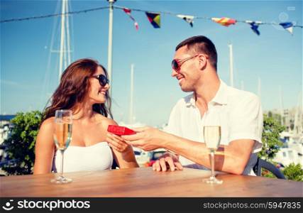 love, dating, people and holidays concept - smiling couple wearing sunglasses with champagne and red gift box looking to each other at cafe