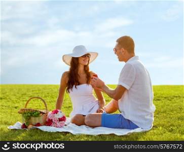 love, dating, people, proposal and holidays concept - smiling young man giving small red gift box with wedding ring to his girlfriend on picnic over blue sky and grass background