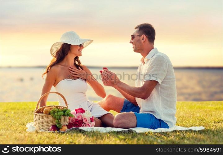love, dating, people, proposal and holidays concept - smiling young man giving small red gift box with wedding ring to his girlfriend on picnic over seaside sunset background