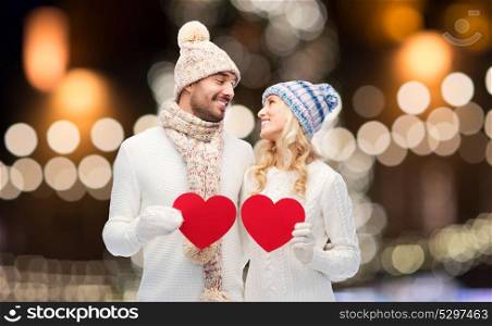 love, holidays and people concept - happy couple in winter hats and scarf holding red paper heart shapes over christmas lights background. couple with red hearts over christmas lights