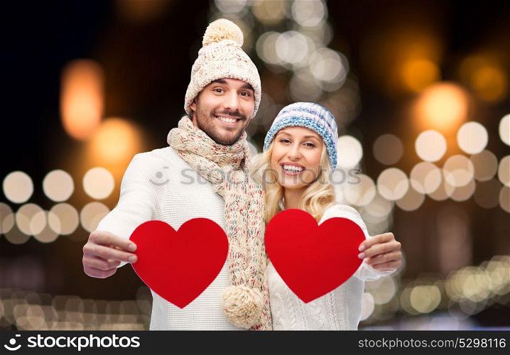love, holidays and people concept - happy couple in winter hats and scarf holding red paper heart shapes over christmas tree lights background. couple with red hearts over christmas lights