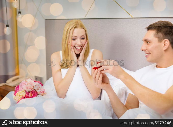 love, holidays, proposal, valentines day and people concept - happy man giving woman red gift box with diamong engagement ring in bed over lights background