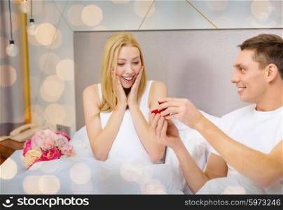love, holidays, proposal, valentines day and people concept - happy man giving woman red gift box with diamong engagement ring in bed over lights background