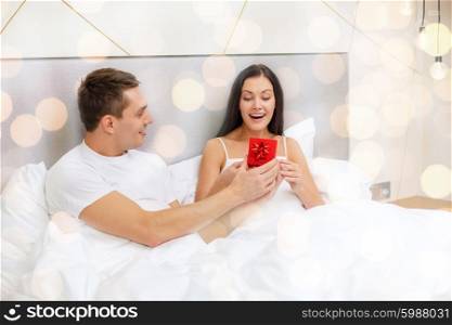 love, holidays, valentines day and people concept - happy man giving woman little red gift box in bed over lights background