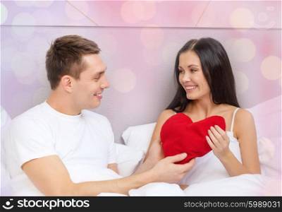 love, holidays, valentines day and people concept - smiling couple in bed with red heart-shaped pillow over holidays lights background