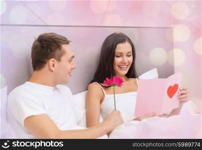 love, holidays, valentines day and people concept - smiling couple in bed with postcard and flower over holidays lights background