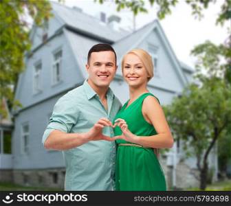 love, people, real estate, home and family concept - smiling couple hugging and showing heart shape gesture over house background