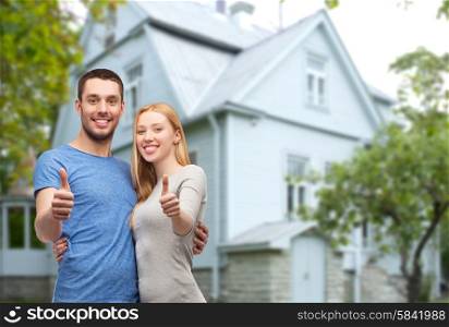 love, people, real estate, home and family concept - smiling couple hugging and showing thumbs up over house background
