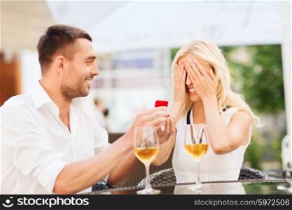 love, proposal, relations, people and holidays concept - happy couple with engagement ring in small red gift box and glasses of wine at restaurant terrace