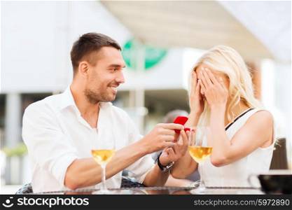 love, proposal, relations, people and holidays concept - happy couple with engagement ring in small red gift box and glasses of wine at restaurant terrace