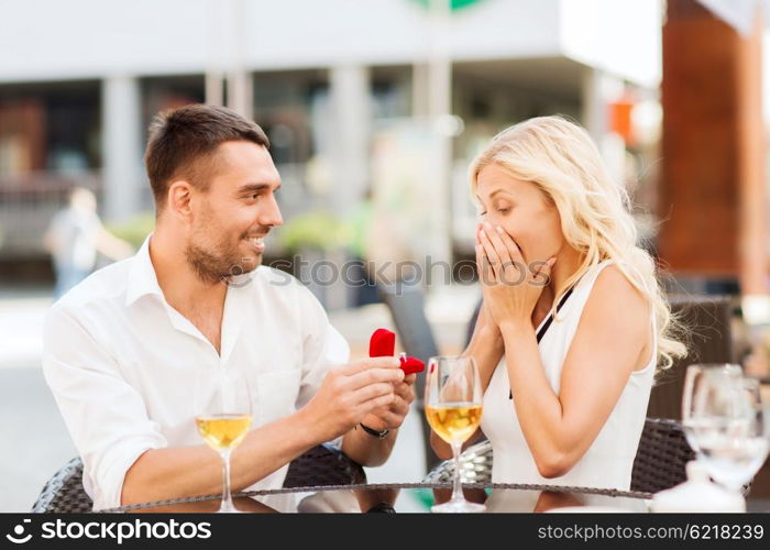 love, proposal, relations, people and holidays concept - happy couple with engagement ring in small red gift box and glasses of wine at restaurant terrace