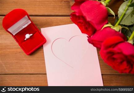 love, proposal, valentines day and holidays concept - close up of gift box with diamond engagement ring, red roses and greeting card on wood