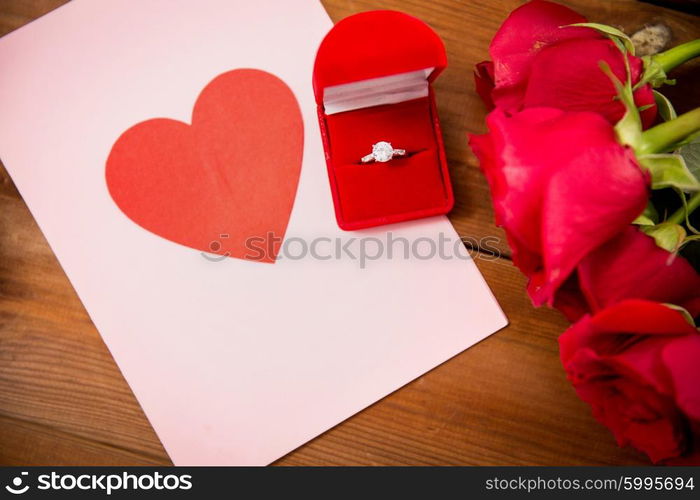 love, proposal, valentines day and holidays concept - close up of gift box with diamond engagement ring, red roses and greeting card on wood