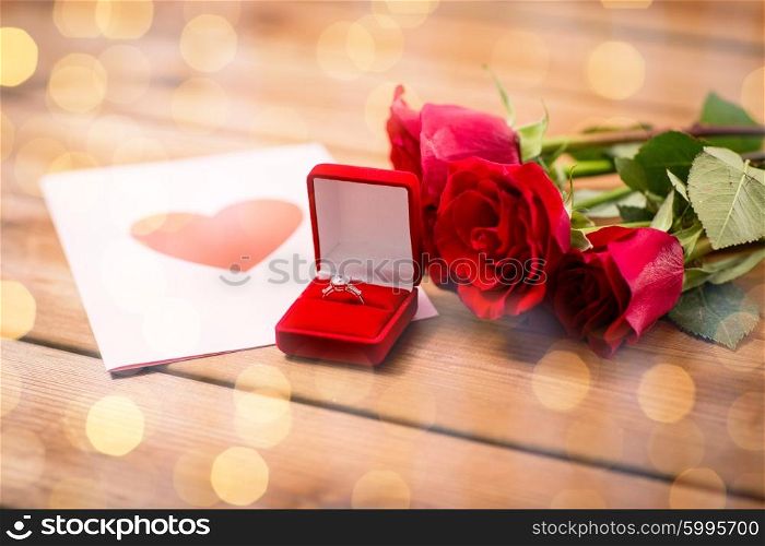 love, proposal, valentines day and holidays concept - close up of gift box with diamond engagement ring, red roses and greeting card on wood