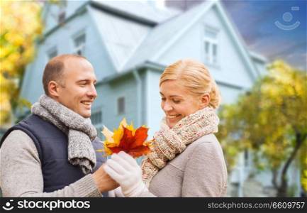 love, relationships and real estate concept - smiling couple with autumn maple leaves over living house background. smiling couple with autumn maple leaves over house