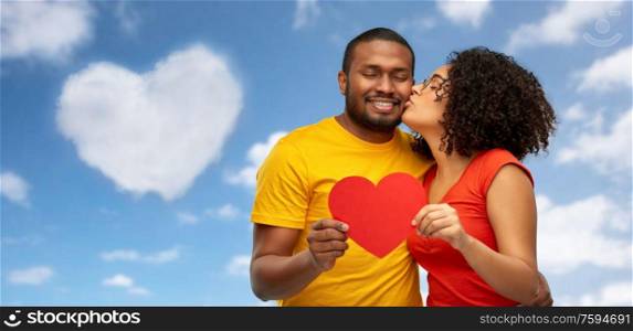 love, relationships and valentines day concept - happy african american couple holding big red paper heart and kiss over blue sky and clouds background. happy african american couple wit red heart kiss