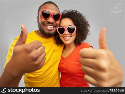 love, relationships and valentines day concept - happy smiling african american couple in heart shaped sunglasses showing thumbs up over grey background. happy african couple in heart shaped sunglasses
